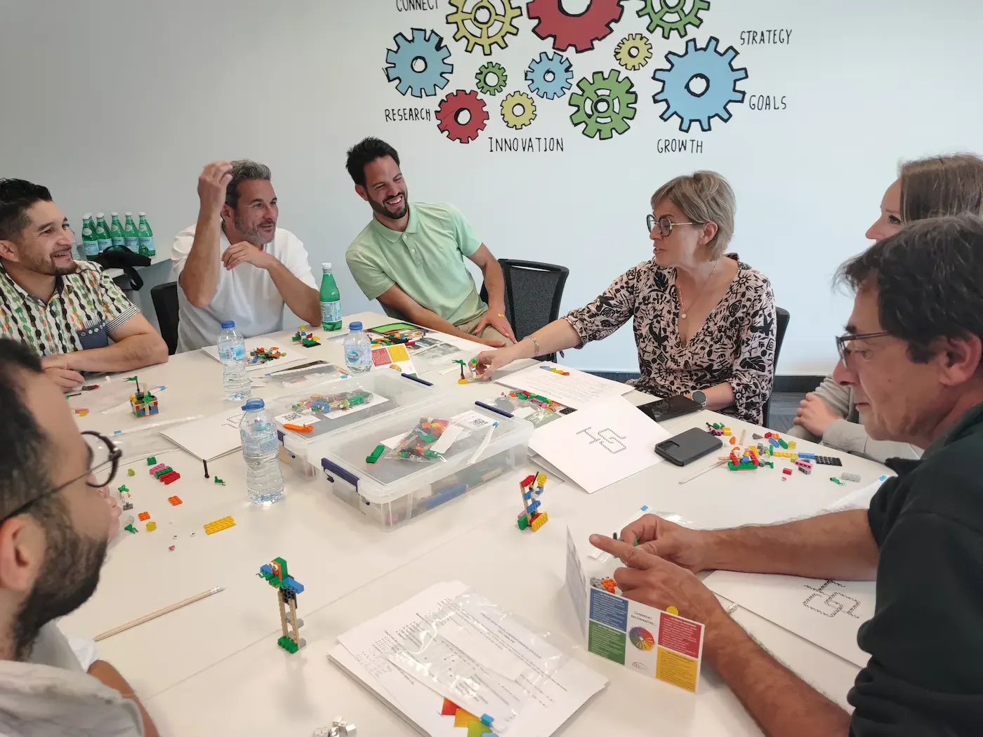 Séminaires 4 20/10/2026 Activité séminaire Premium : une équipe échange et assemble des briques LEGO® autour d'une table, devant un mur décoré d'engrenages