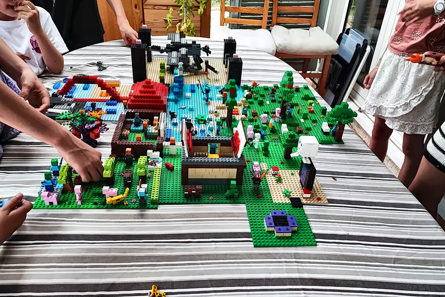 Enfants réunis autour d'une table à la maison pendant un anniversaire à domicile : ils construisent ensemble un village en briques LEGO® sur des plaques vertes