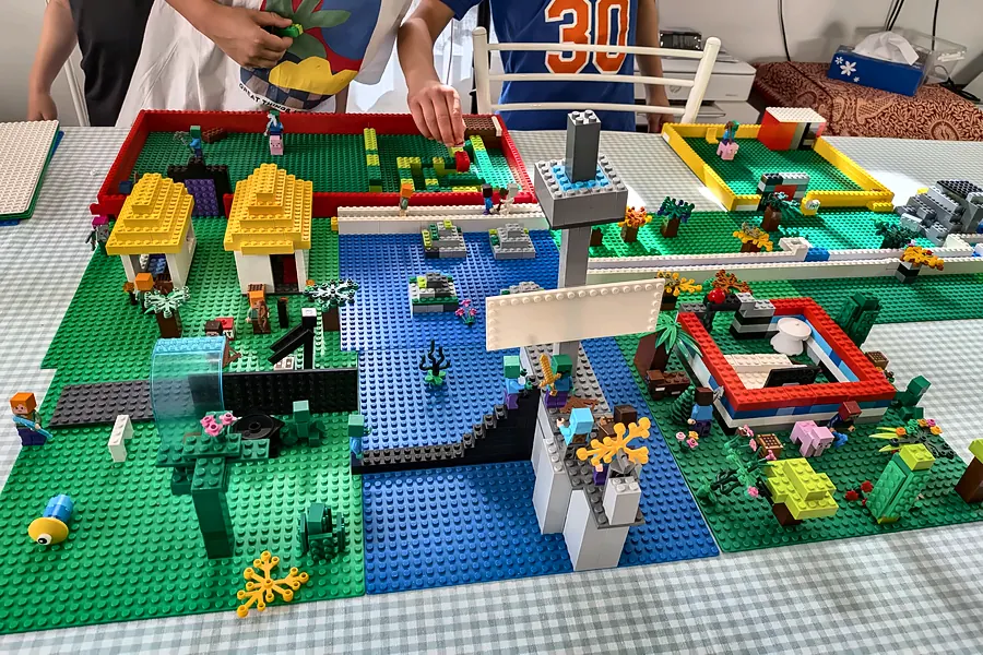 Village avec rivière et maisons jaunes en briques LEGO®, construit sur une table pendant un anniversaire à domicile, pendant que les enfants posent leurs briques