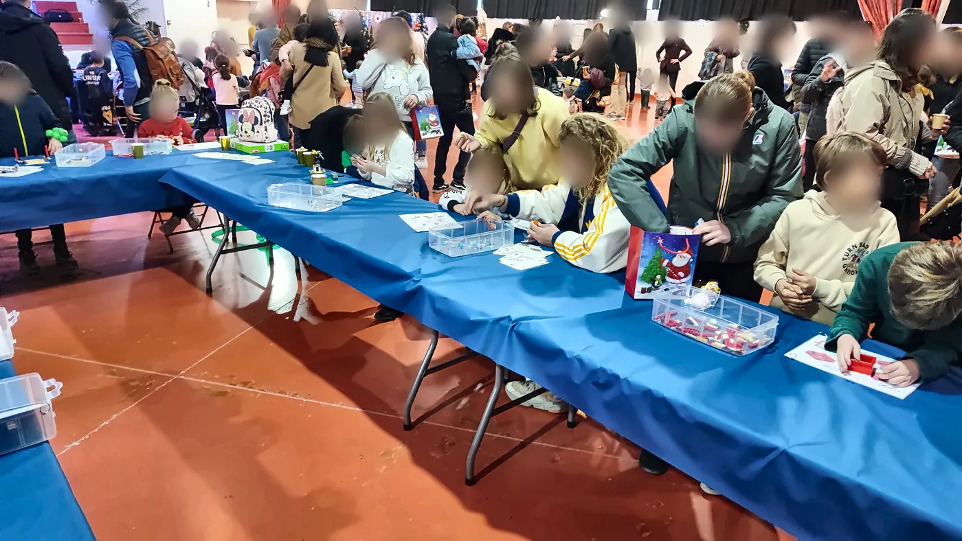 Arbre de Noël entreprise : enfants et parents construisent côte à côte sur des tables nappées de bleu, avec une notice et un sac cadeau de Père Noël