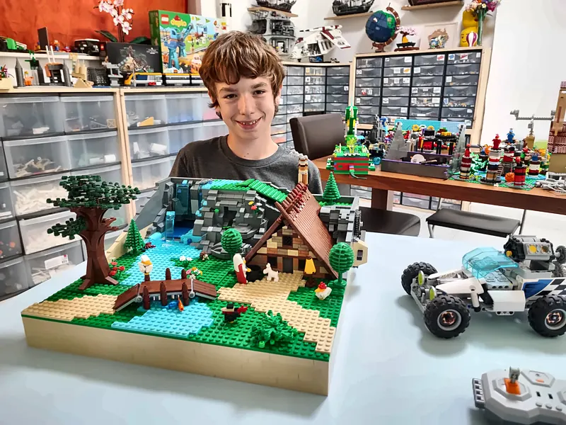 Un participant souriant montre son diorama de la hutte de Panoramix, dans Astérix®, en briques LEGO® : maison de bois, grand arbre et cascade motorisée dont de petites briques s'écoulent en circuit fermé