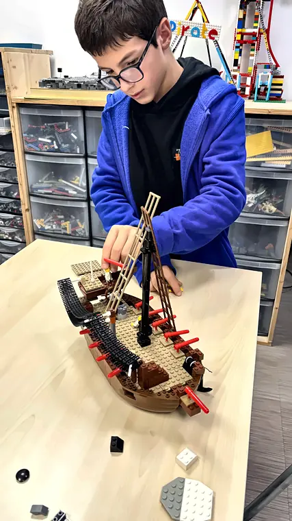 Accès libre 11 20/10/2026 Un jeune garçon assemble les voiles d'un galion pirate en briques LEGO® en accès libre, devant les tiroirs de pièces