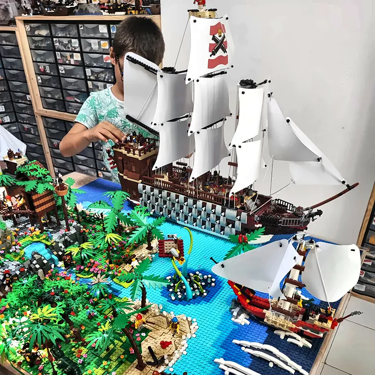 Stage vacances 9 20/10/2026 Participant d'un stage vacances scolaires qui pose les voiles d'un vaisseau de ligne pirate en briques LEGO®, au milieu d'une île aux palmiers et de petits voiliers