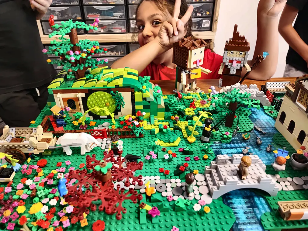 Stage vacances 15 20/10/2026 Hobbiton, dans Le Seigneur des Anneaux®, en briques LEGO® : jardin fleuri, maison de hobbit à porte ronde, pont de pierre et ruisseau, avec une participante qui fait le signe de la victoire