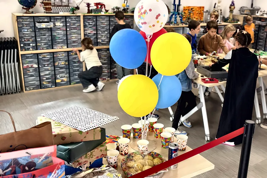 Table de goûter d'anniversaire avec ballons bleus et jaunes, gâteaux et gobelets, dans l'espace créatif de Salon-de-Provence, pendant que les enfants construisent en briques LEGO® au fond de la salle