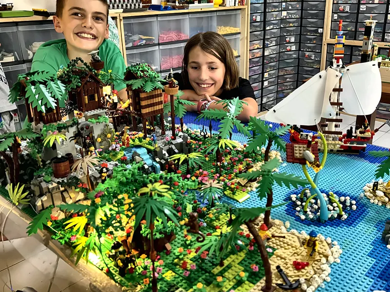Deux participants souriants penchés sur une île de pirates en briques LEGO® avec palmiers, cabanes et voiliers
