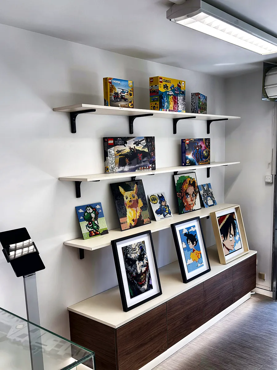 Accueil 15 20/10/2026 Tableaux en pièces LEGO® encadrés et boîtes exposés sur des étagères de la boutique